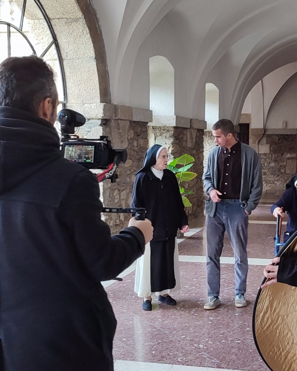 Un cámara del equipo graba a una monja y a un joven que conversan en el claustro de un monasterio.